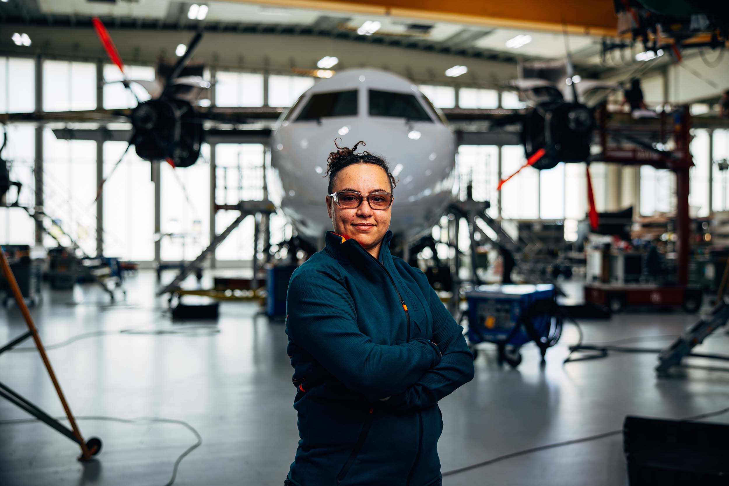 Eine Frau mit Brille und schwarzen Haaren steht mit verschränkten Armen vor einem Weißen Propeller Flugzeug in einer Halle. Die Frau trägt eine grüne Jacke.