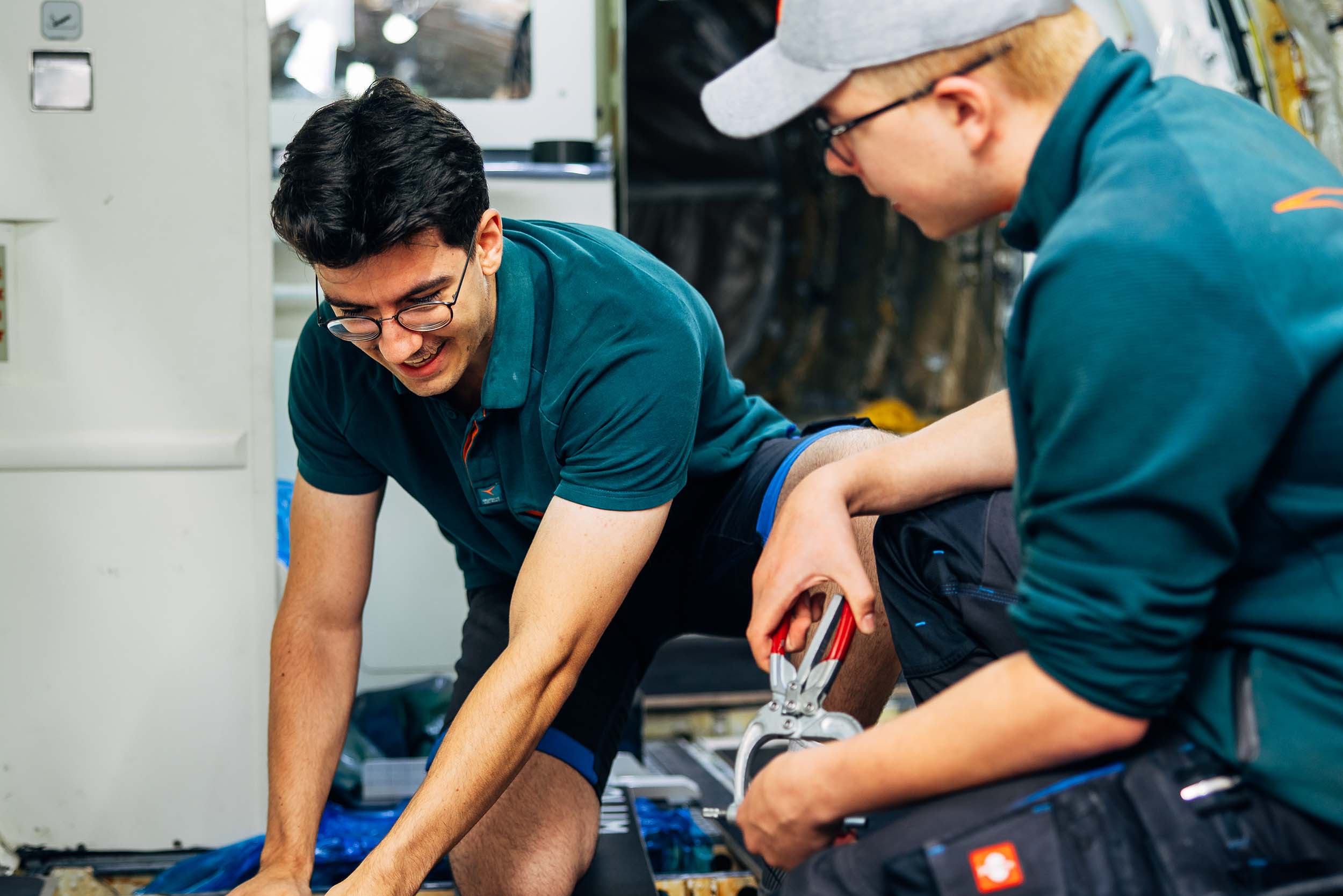Zwei junge Männer arbeiten gemeinsam an einem Projekt. Ein Mann hat schwarze kurze Haare und eine Brille. Der andere Mann hat eine Zange in der Hand und hat eine graue Kappe auf dem Kopf. Beide tragen ein grünes Polo Shirt.