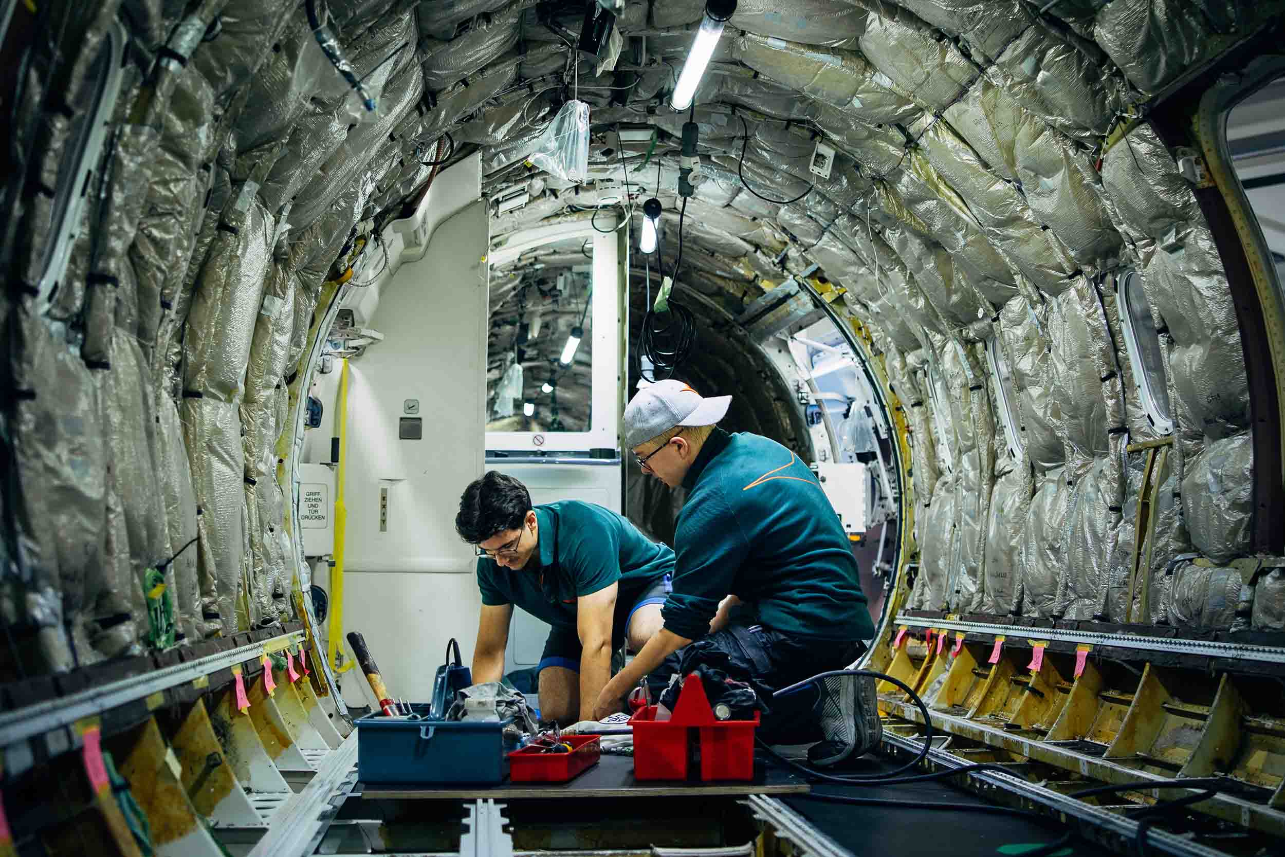 Zwei Männer sind in einer Flugzeugkabine. Die Kabine ist entkernt, dies erkennt man durch Lampen und Dichtungsmaterial an der Wand. Die Männer arbeiten am Boden, neben ihnen steht eine Werkzeugkiste.